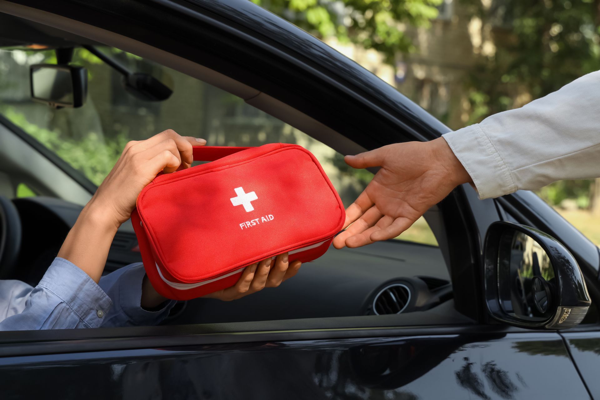 Person hands a red first aid kit through a car window on a sunny street