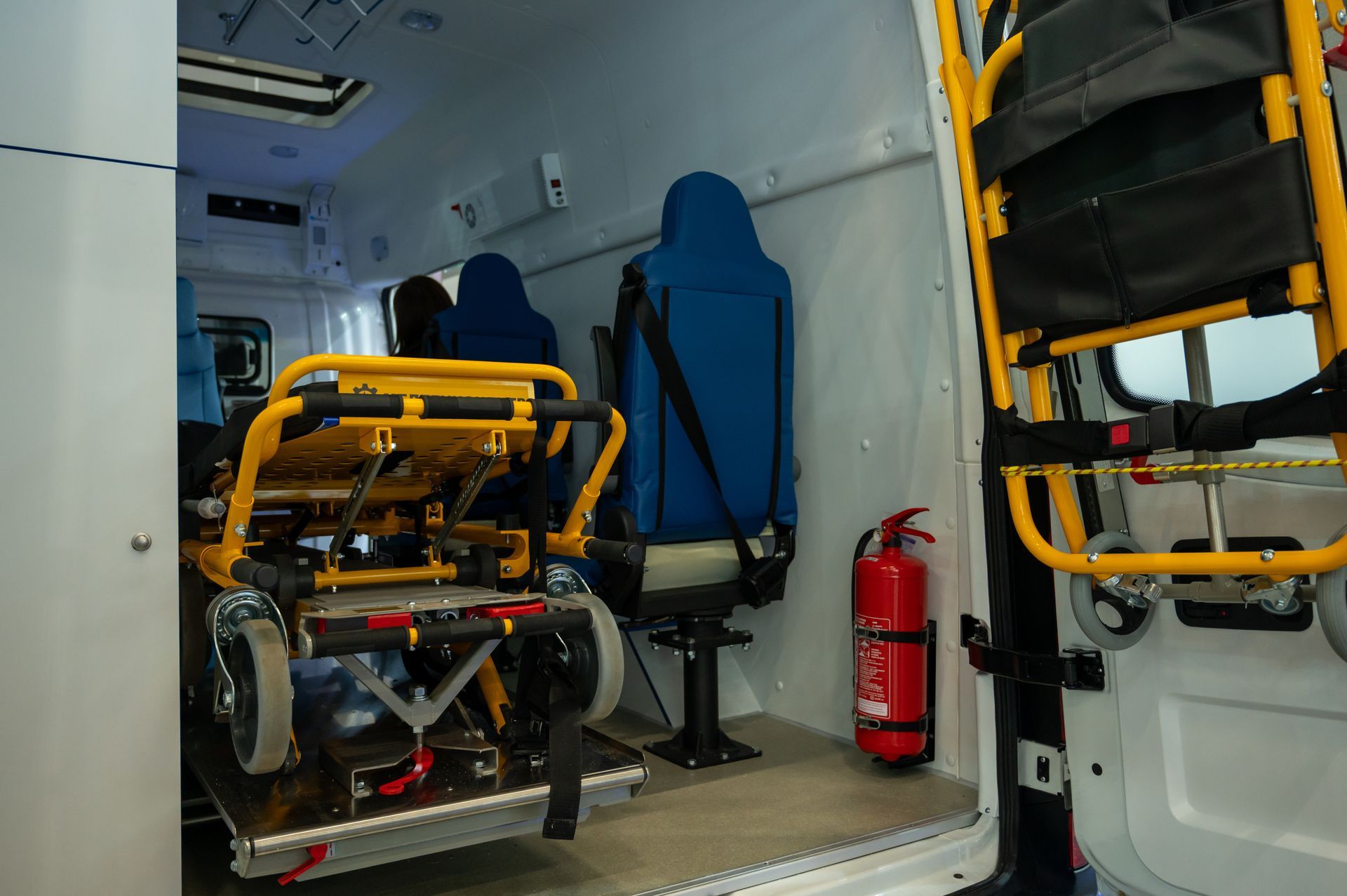 Interior of an ambulance with a wheelchair secured inside, blue seat and red fire extinguisher visible.