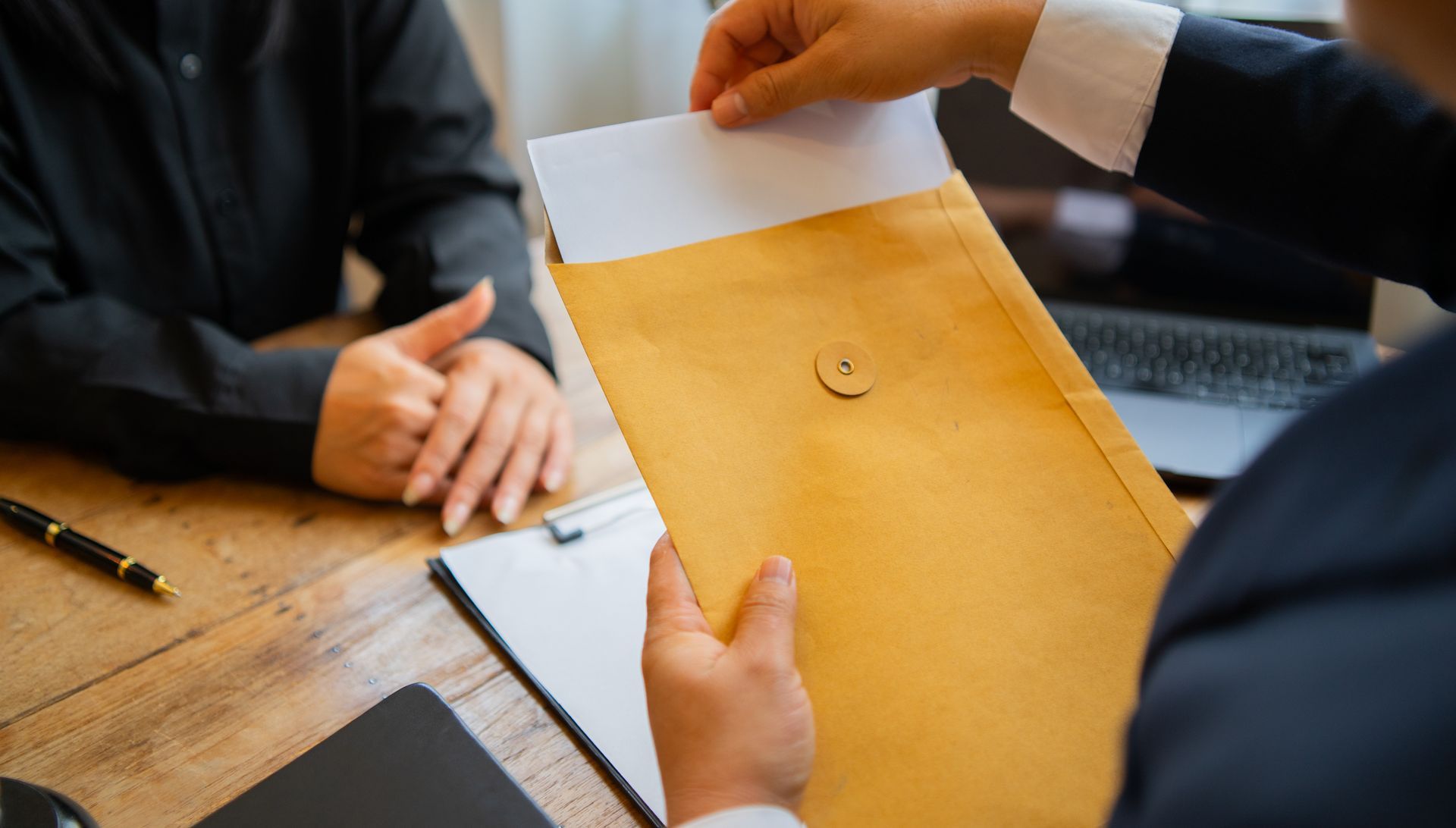Hands exchanging a manila envelope over a wooden desk in an office meeting