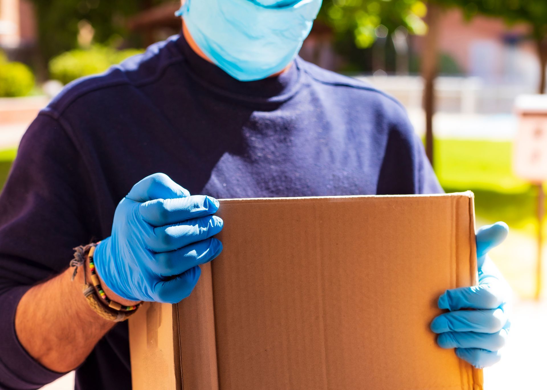 Masked delivery worker holding a cardboard package outdoors, wearing blue gloves