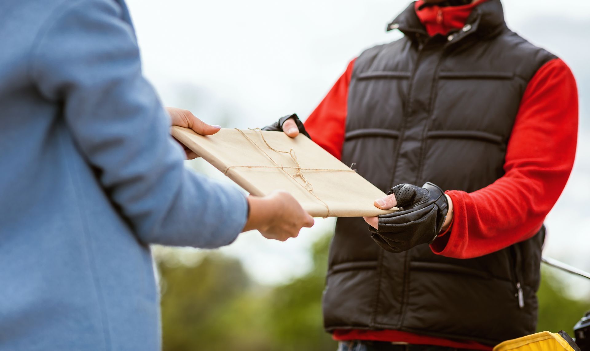 Delivery person in red jacket handing a cardboard package to someone in a blue coat outdoors
