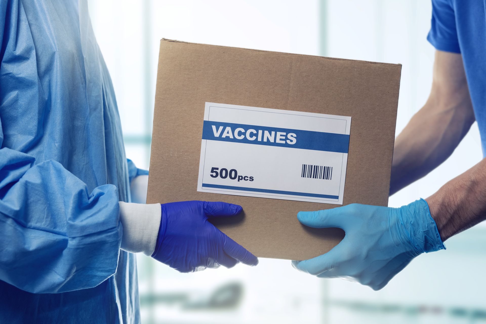 Gloved hands handing a cardboard box labeled Vaccines between two people