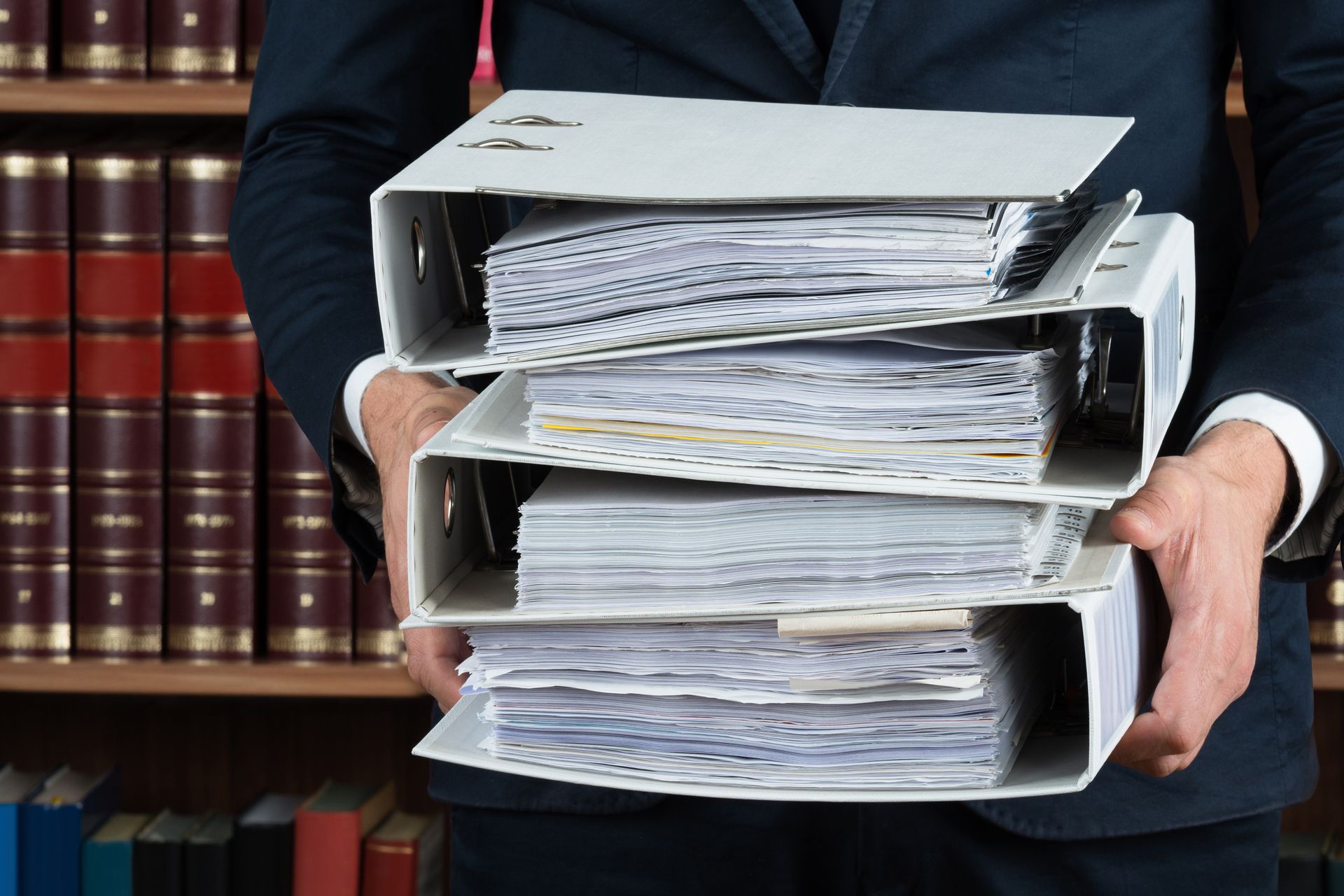 Person holding a stack of thick file binders in front of a bookshelf