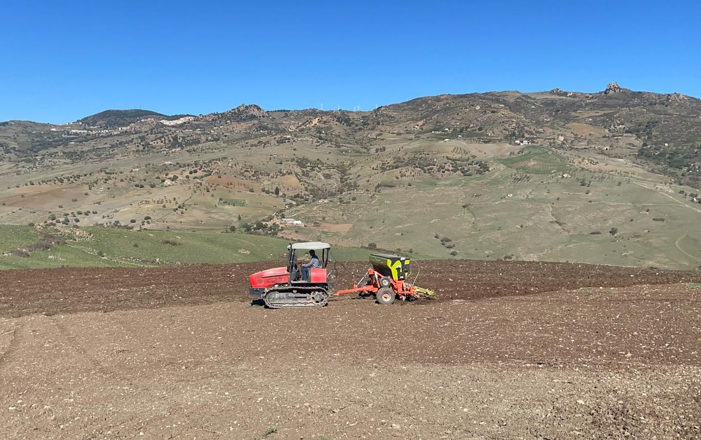 Un trattore cingolato rosso traina attrezzi agricoli attraverso un campo arato, sullo sfondo di montagne ondulate e aride.