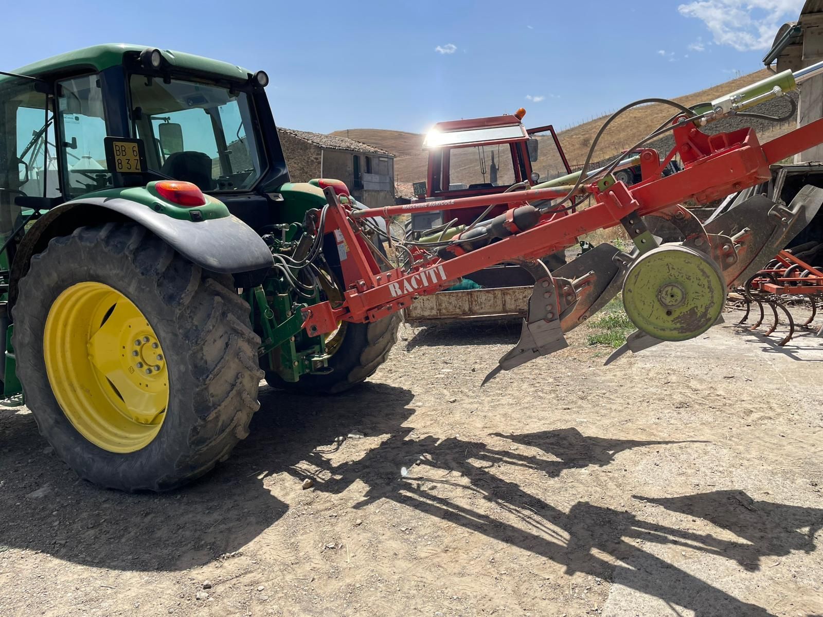 Un trattore John Deere verde con un aratro agricolo rosso, parcheggiato su un terreno sterrato sotto un cielo azzurro e soleggiato.