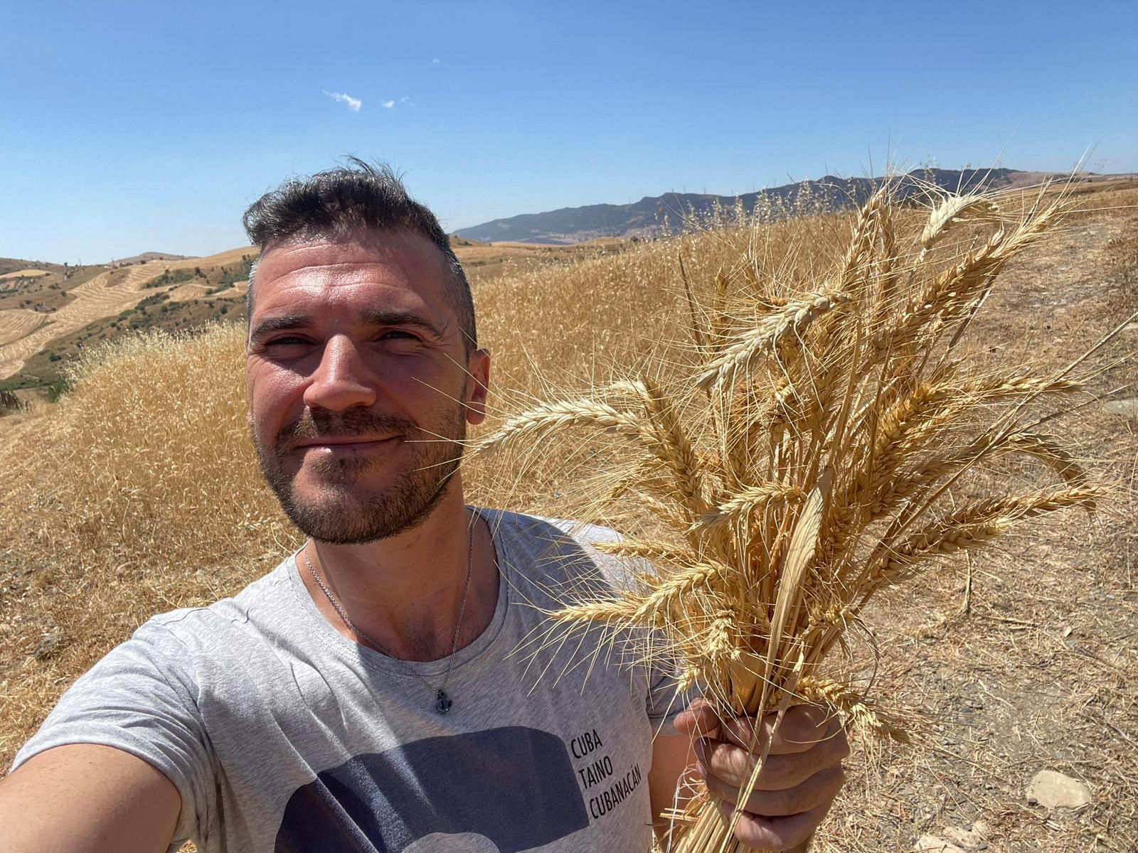Una persona sorride mentre tiene in mano un grosso fascio di grano dorato essiccato in un vasto, soleggiato e arido paesaggio collinare.