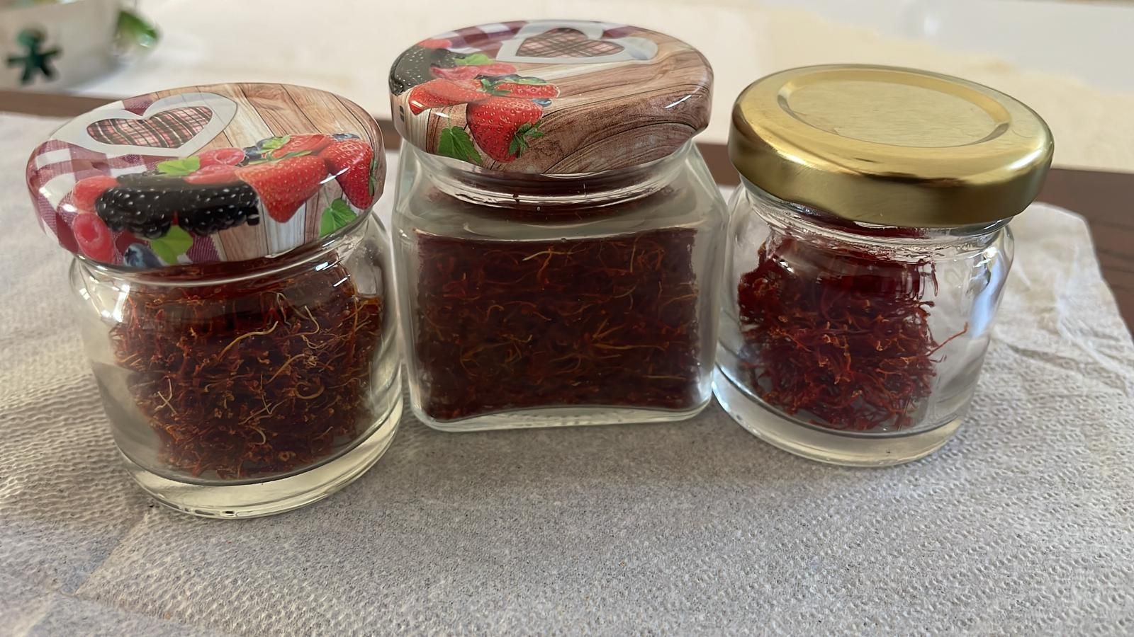Tre piccoli vasetti di vetro pieni di fili di zafferano rosso, appoggiati su una superficie di stoffa chiara.