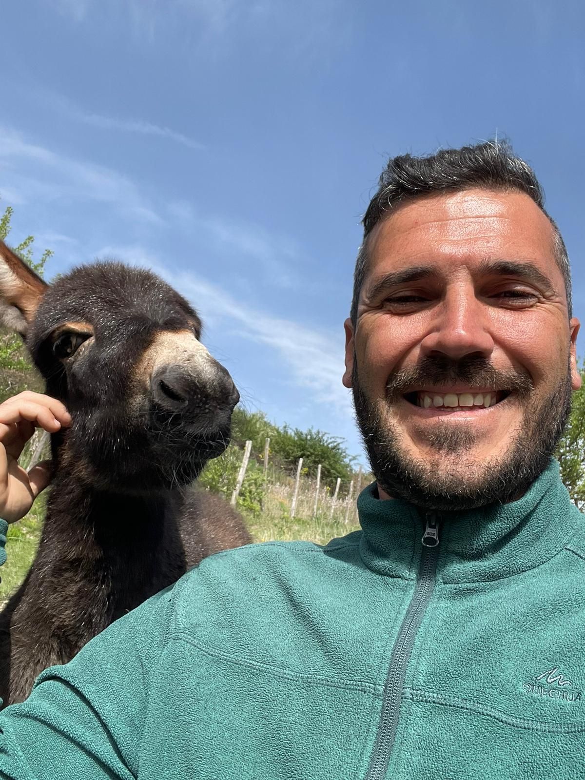 Un uomo con indosso un pile verde sorride mentre accarezza un piccolo asinello scuro, sullo sfondo di un cielo azzurro e limpido.