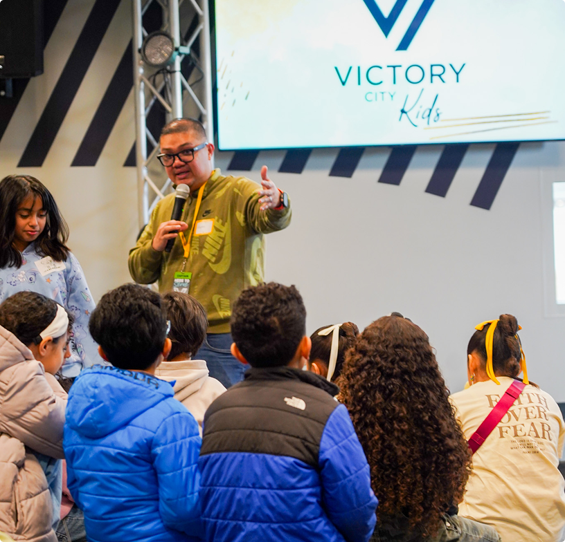 Speaker addressing seated audience at Victory City Kids event, holding microphone on stage