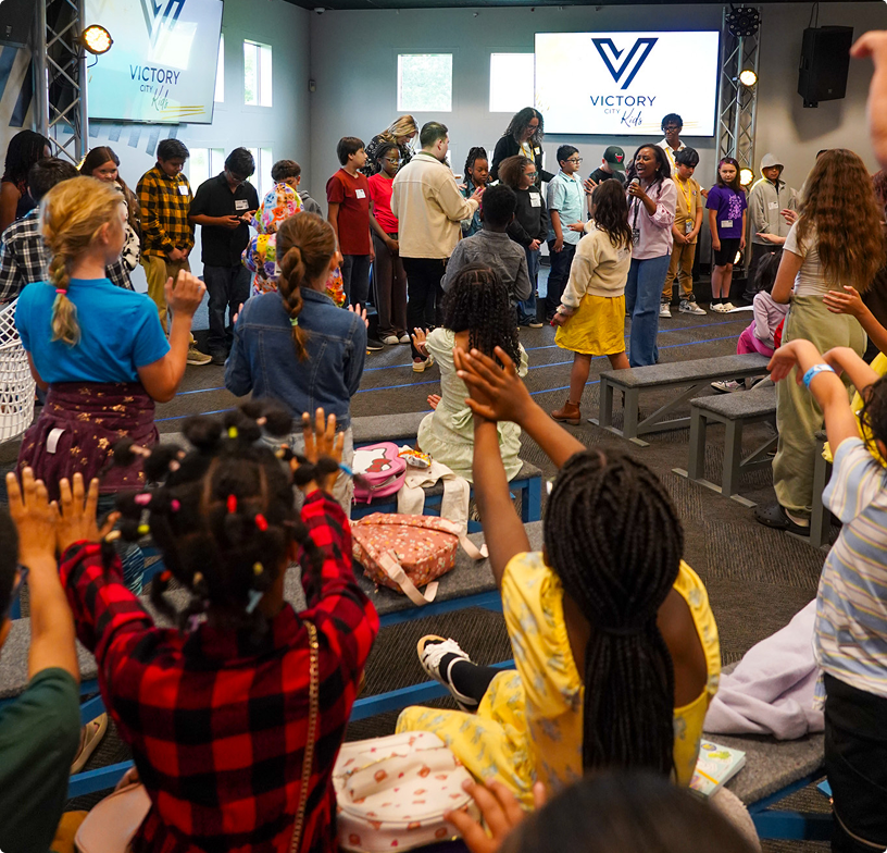 Crowded indoor worship gathering with people seated and standing facing a stage and “Victory” screen