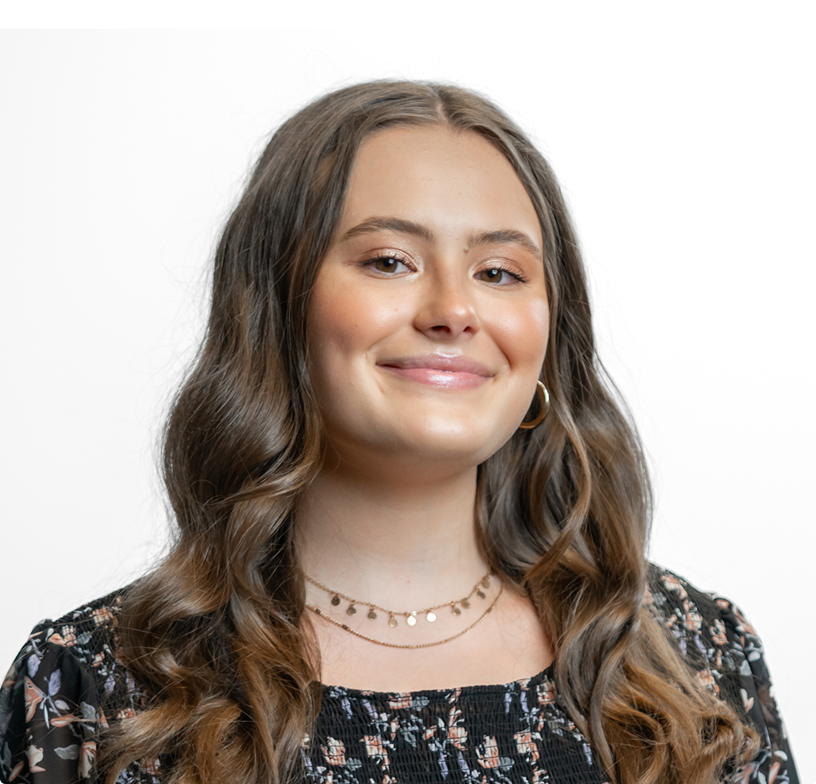 Smiling woman with wavy brown hair wearing a floral dress and layered necklace against a white background