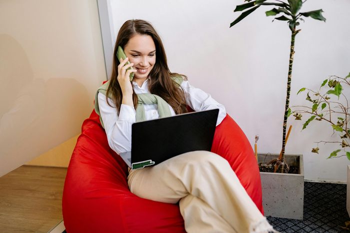 Woman on red beanbag chair, talking on the phone while using a laptop. Bright office setting with a plant.