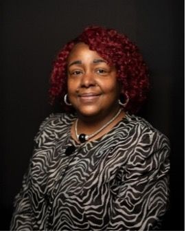 Woman with glasses, arms crossed, wearing a black blazer and dress, smiling.