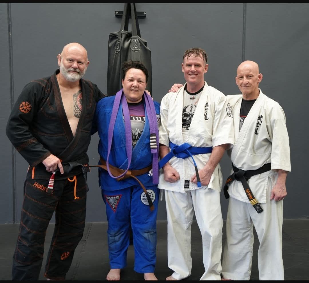Four people in martial arts attire pose together. Two in white gis, one in a black gi, one in a blue gi with a purple belt.