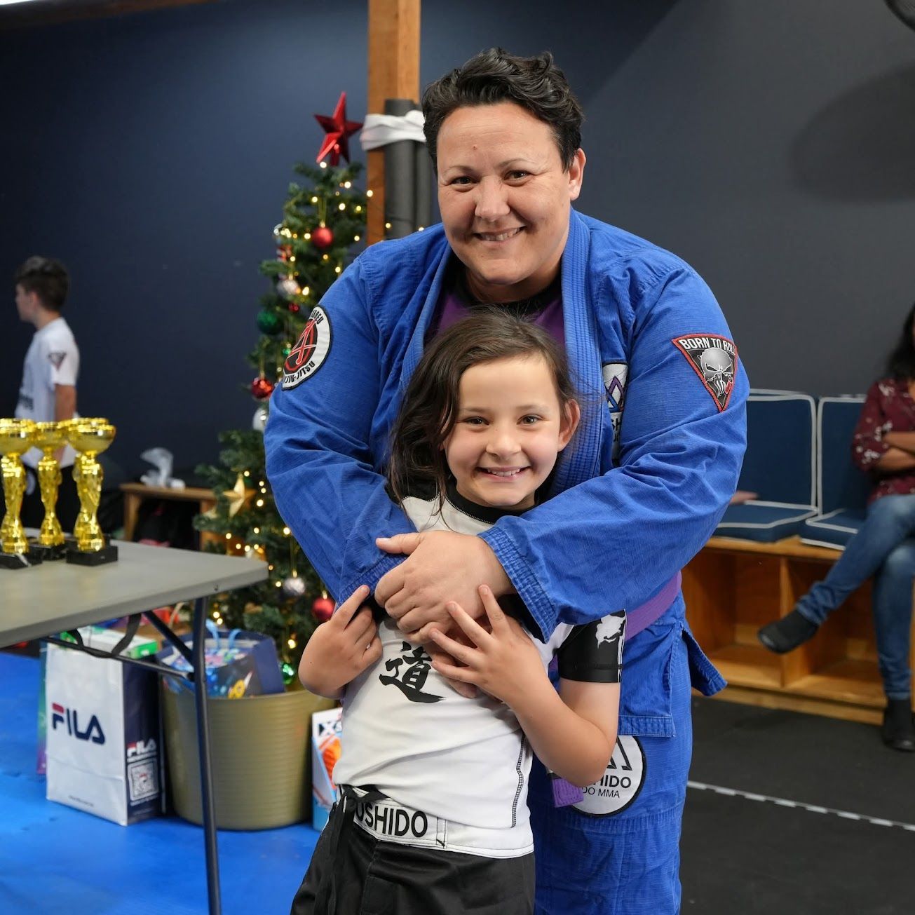 Person in blue gi hugs a child, both smiling, near a Christmas tree and trophies.