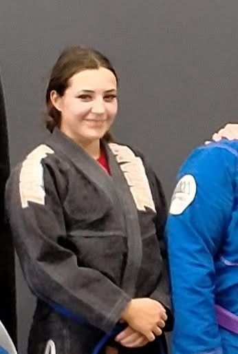 Woman in dark gi smiles, standing next to a person in a blue gi.