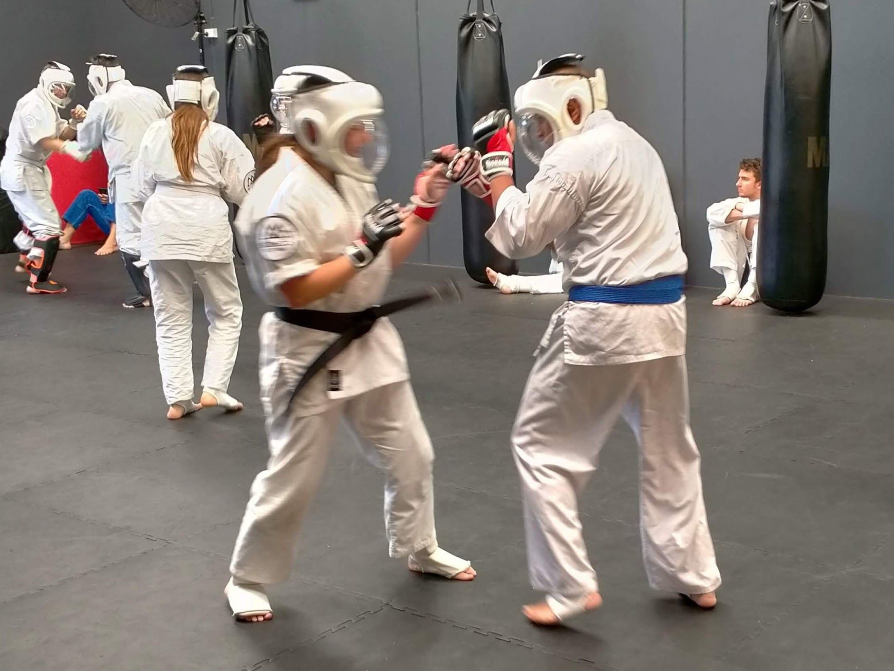 Two people in white karate uniforms sparring in a dojo, wearing protective headgear and gloves. Others practice in the background.