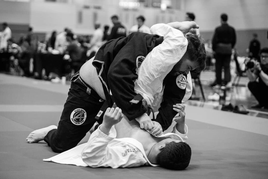 Two people grappling in a Jiu-Jitsu match on a mat, one in a black gi, one in a white gi.