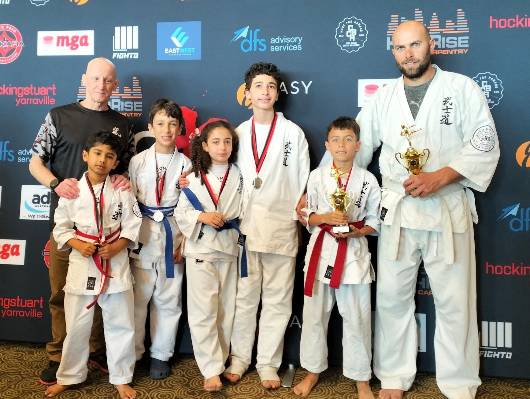 Karate competitors with medals and trophies posing with coaches. They are standing in front of a backdrop with logos.