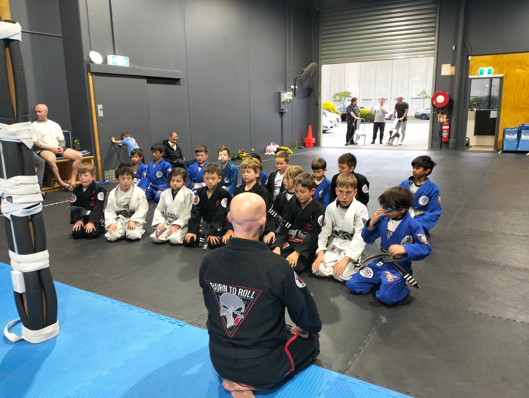 Children in martial arts uniforms kneeling, listening to a coach in a gym.