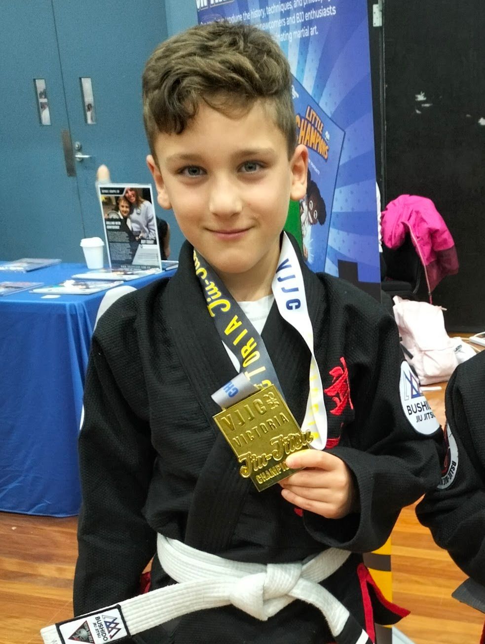 Boy in a black gi holding a gold medal, smiling. Indoor setting with a banner.