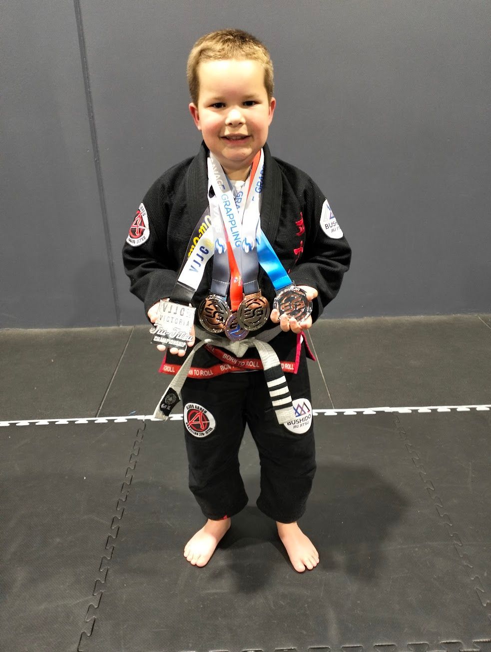 Boy in black gi with medals smiles, standing on mat.