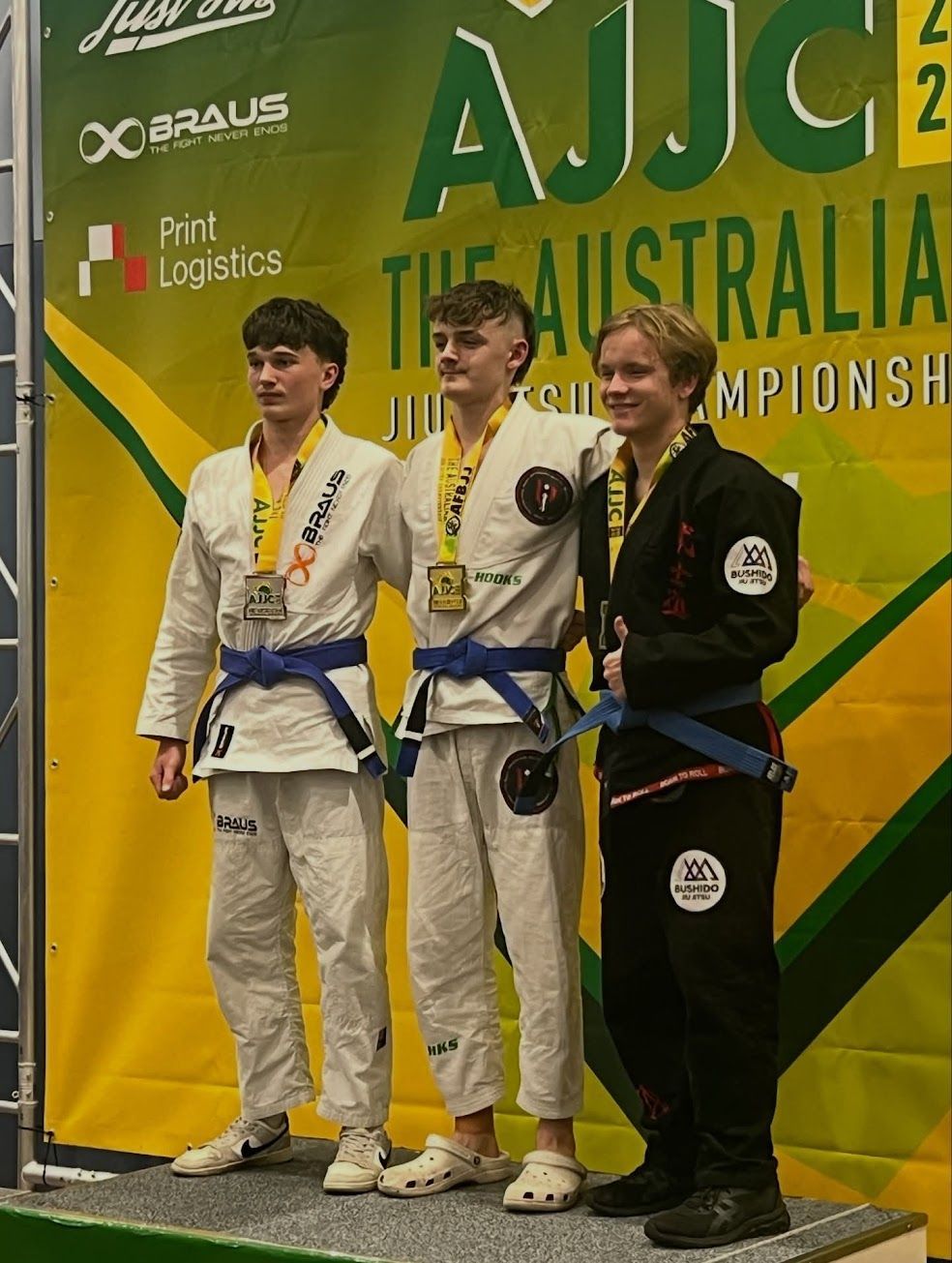 Three athletes on a podium, wearing Jiu-Jitsu gis with medals. Australia Jiu-Jitsu Championship background.