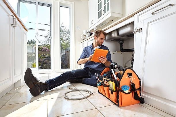 Technician kneeling on kitchen floor beside orange tool bag, inspecting plumbing under the sink.
