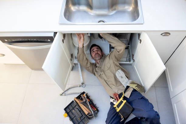 Plumber working under a sink, reaching into plumbing with tools laid out on the floor.