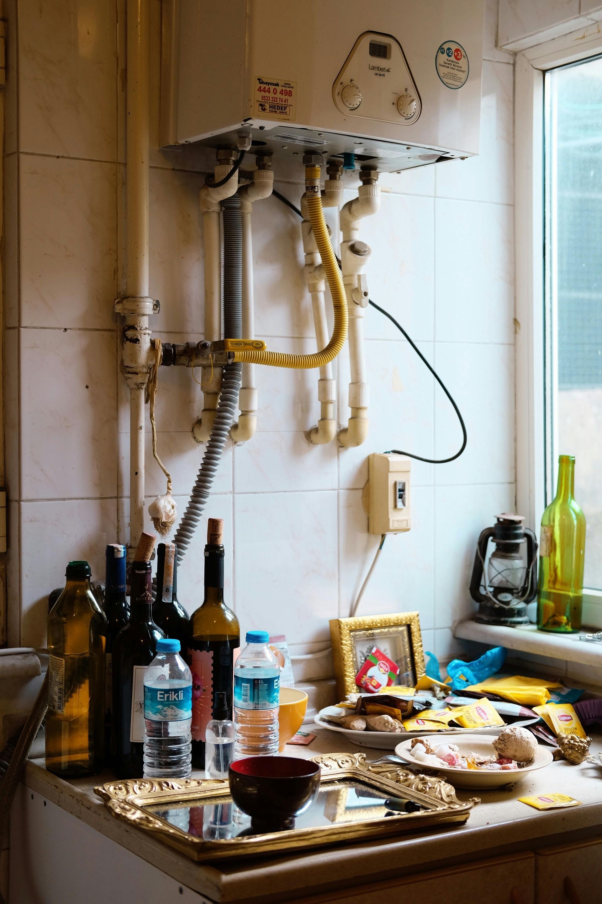 Cluttered kitchen counter with bottles, dishes, food, and a wall-mounted water heater by a window