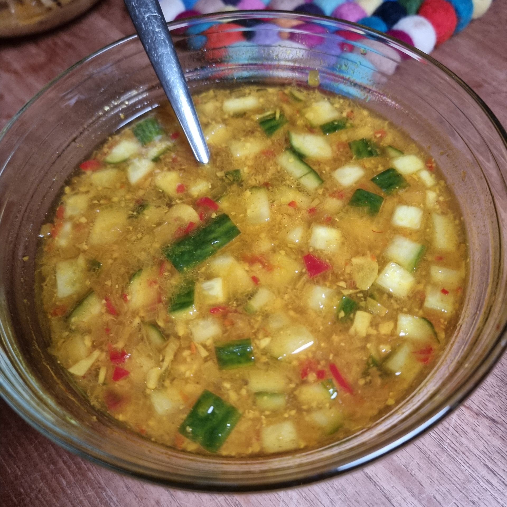 Glazen kom met geelbruine saus, blokjes komkommer en rode paprika. Een zilveren lepel ligt in de kom.