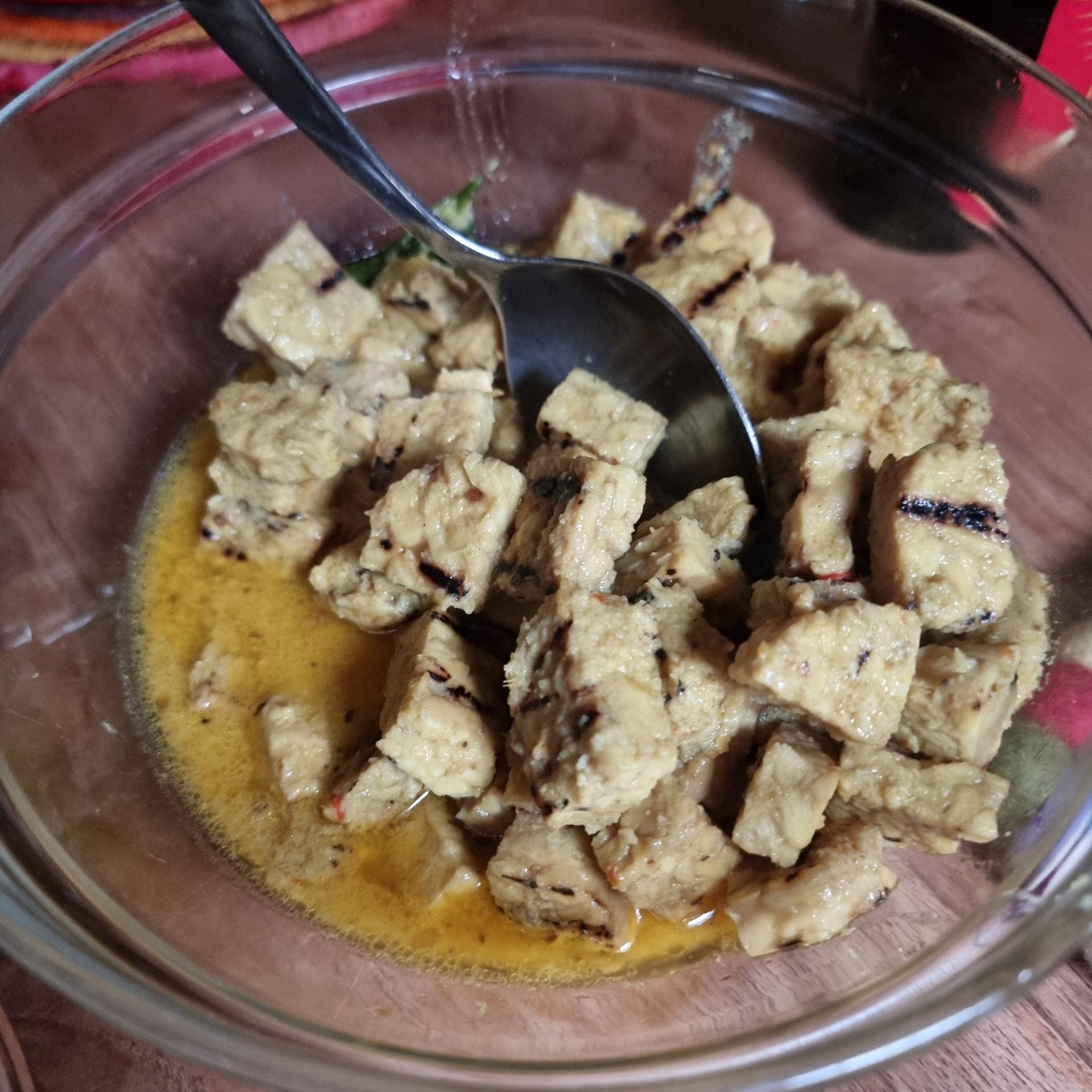 In een doorzichtige kom gesneden tofu met saus, geserveerd met een lepel. Sommige tofu heeft grillstrepen.