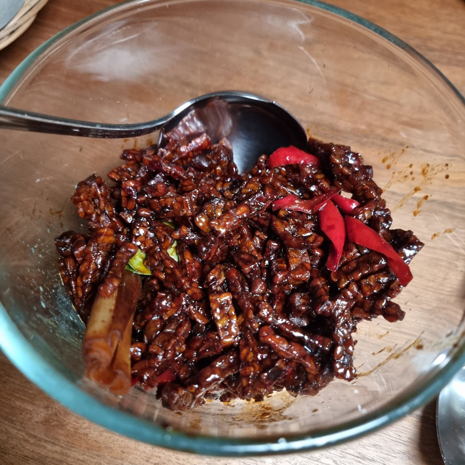 Een schaaltje tempeh in een glazen kom, donkerbruin met rode chilipepers en een lepel op een houten ondergrond.