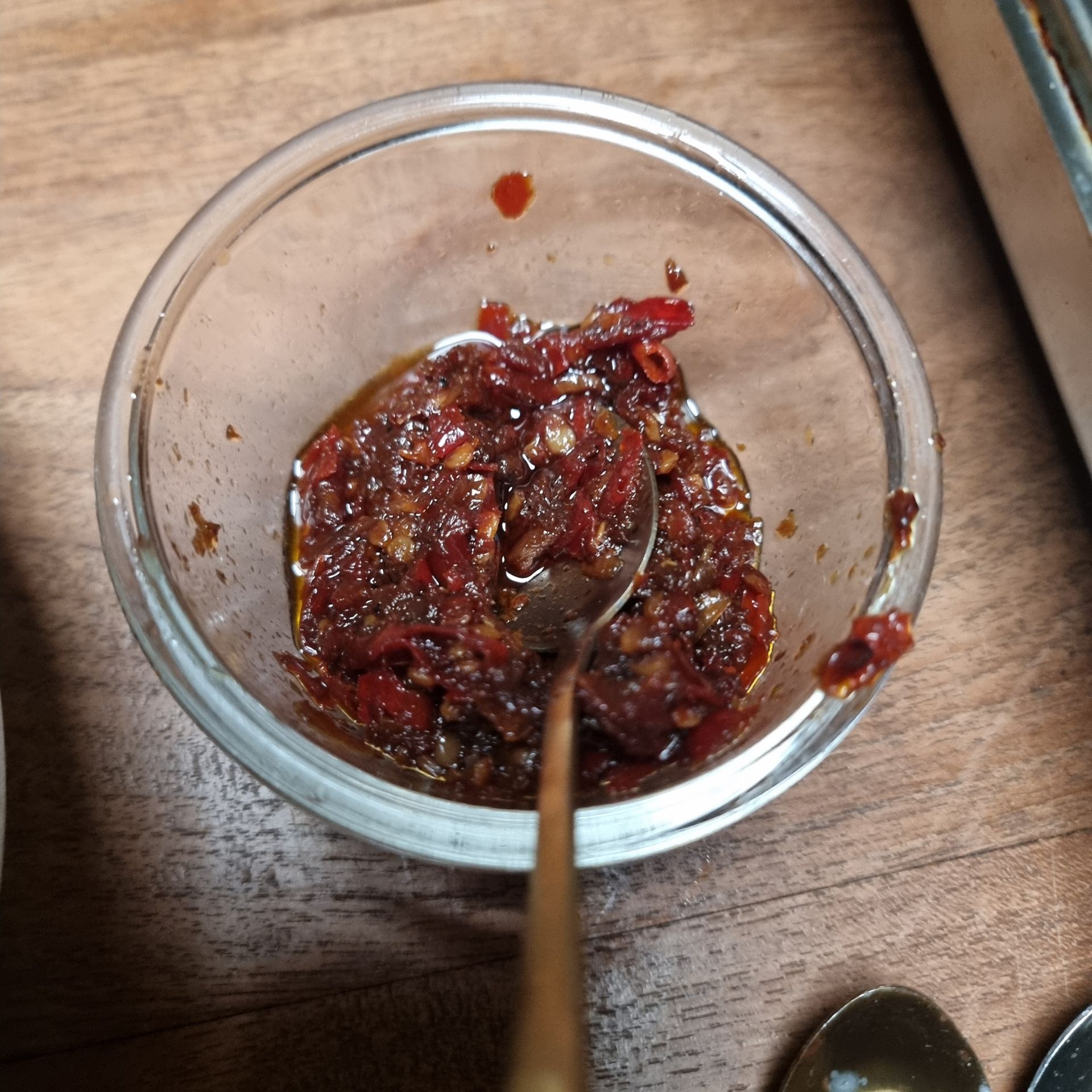 Een klein glazen kommetje met rode chili-olie en een lepel op een houten ondergrond.