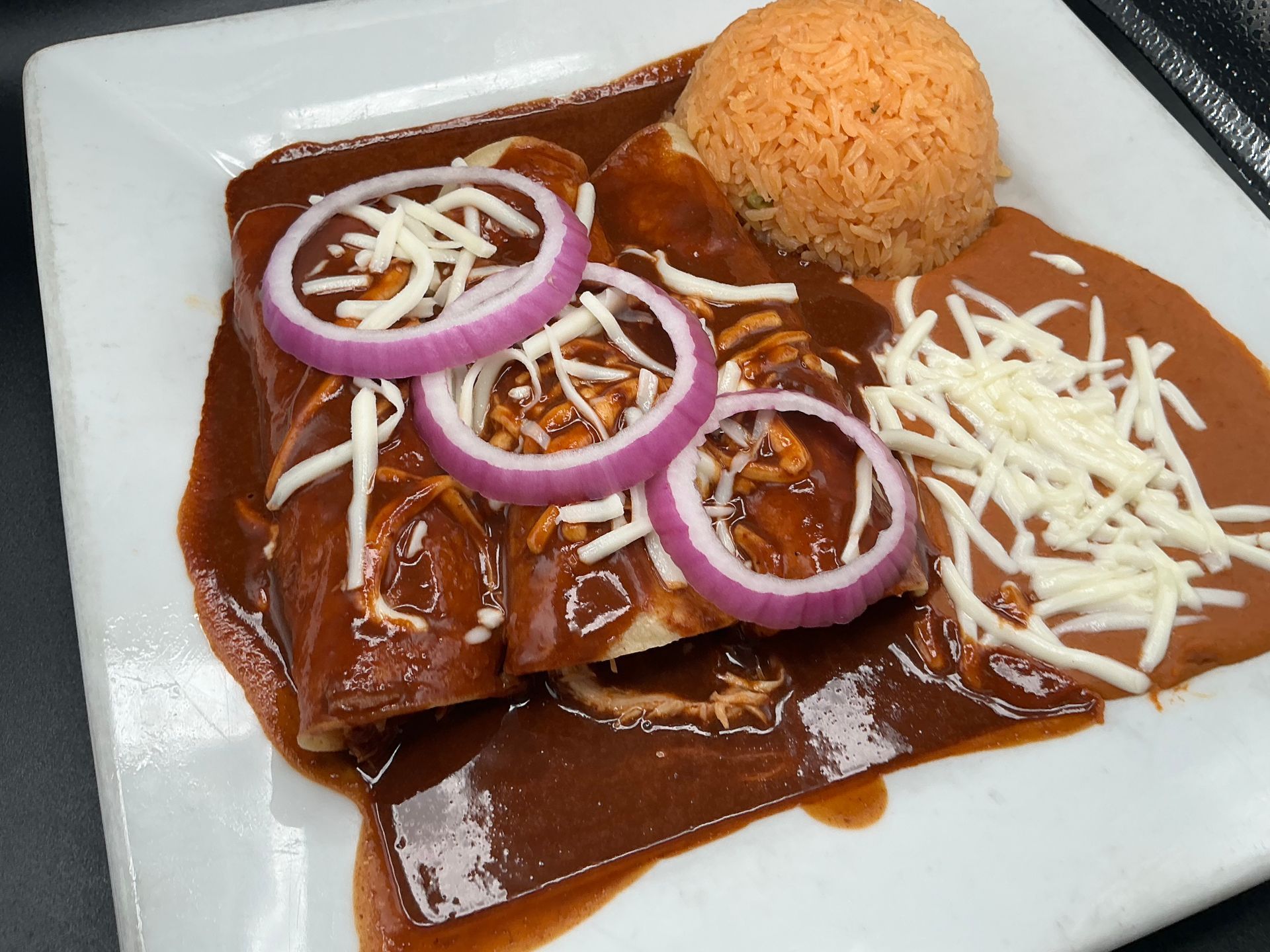 Enchiladas in dark sauce, topped with onion and cheese, with rice on a white plate.