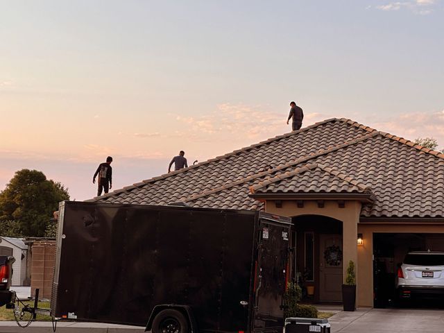 Three people on a roof working; black trailer in front of a house at sunset.