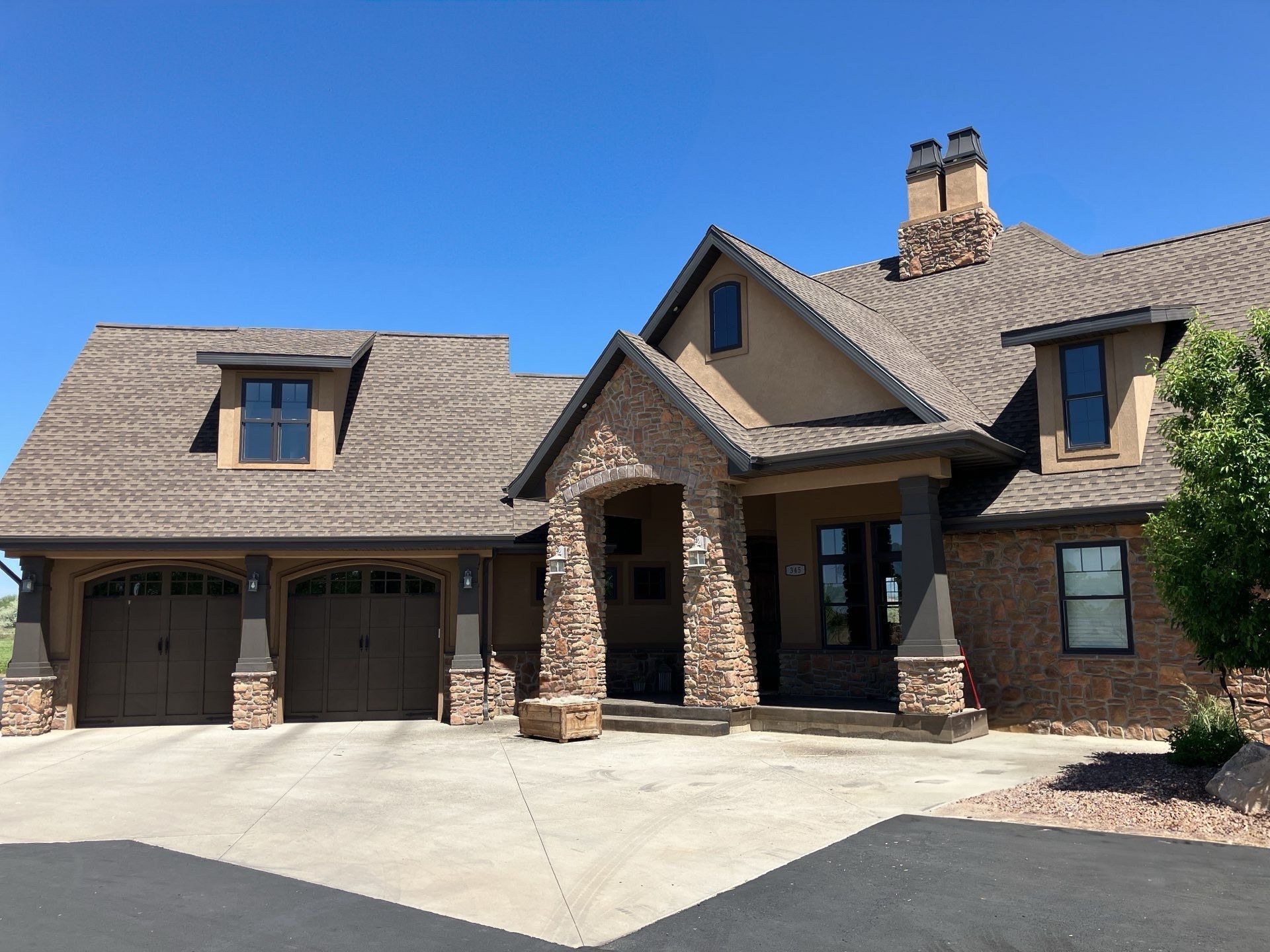 A large, tan and stone house with a brown roof and garage doors on a sunny day.