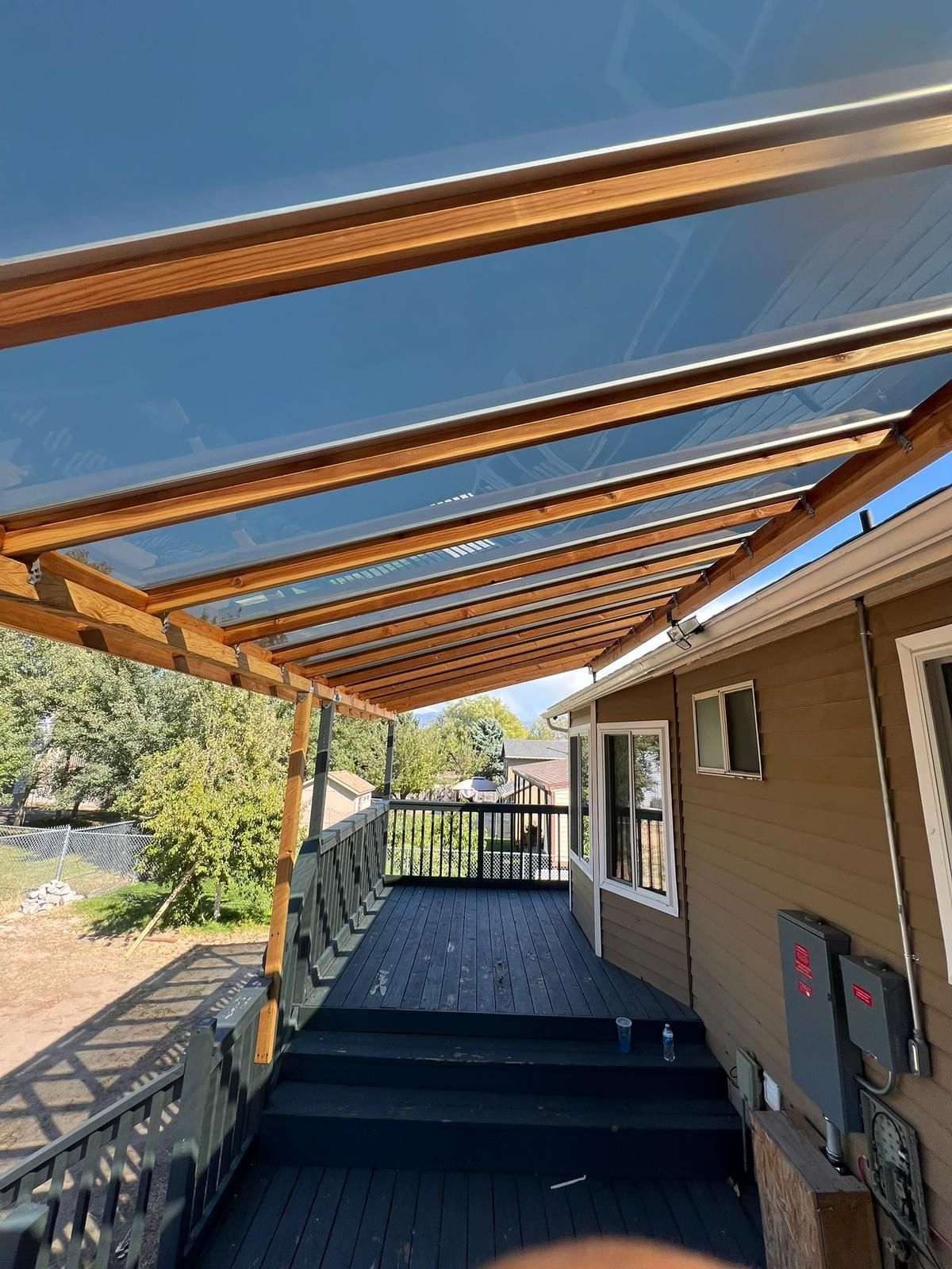 Wooden deck with a clear-roofed patio cover. Overlooks trees and a house with brown siding.