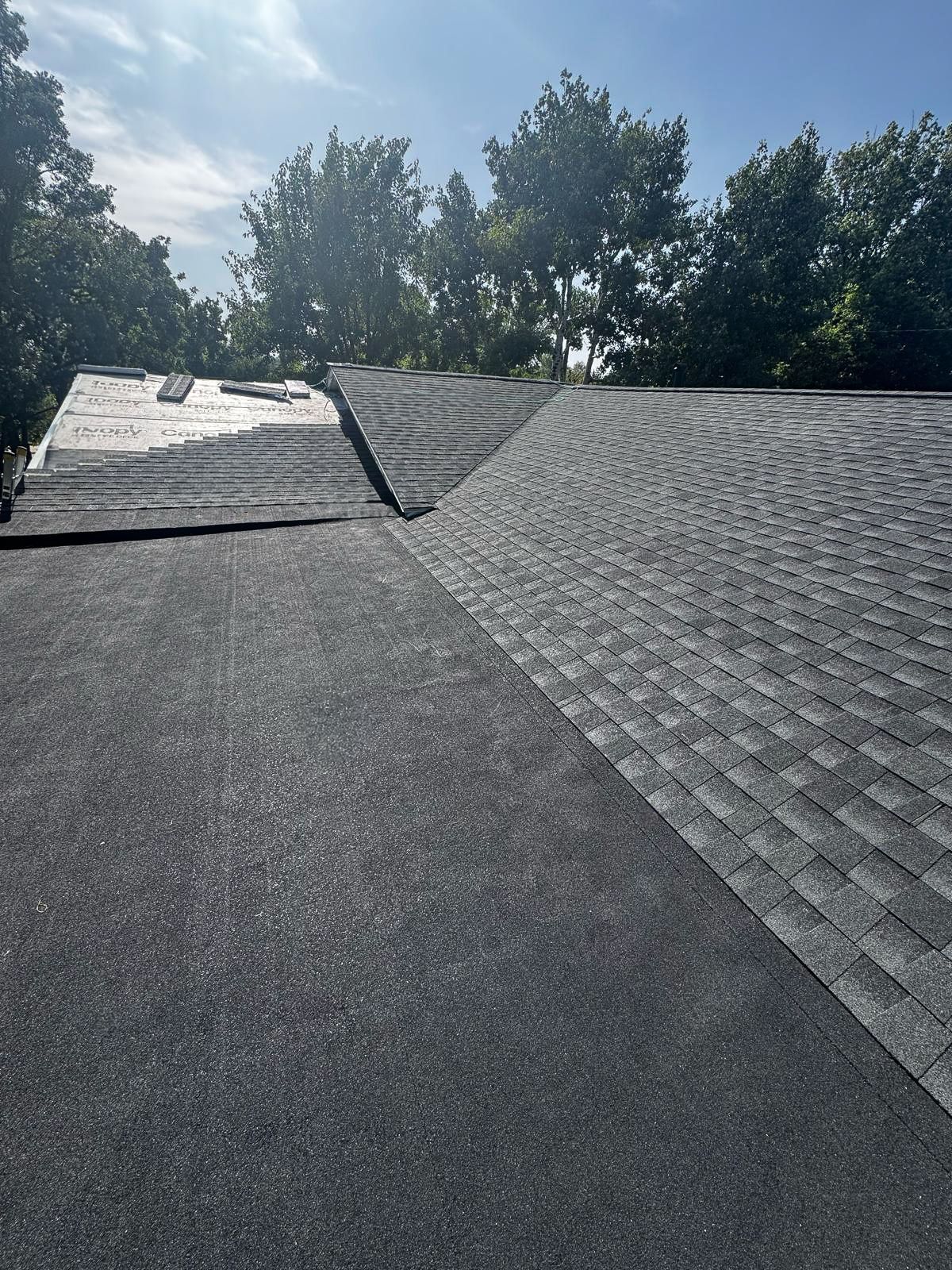 View of a roof with two different types of dark roofing material, trees in the background, and a bright sky.