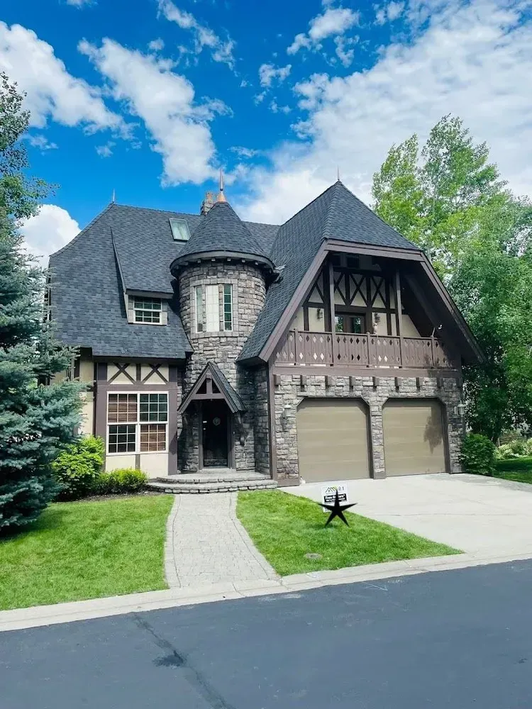 Stone and wood Tudor-style house with arched roof and two-car garage under a blue sky.