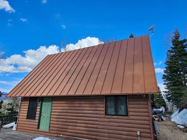Cabin with brown metal roof, log siding, green door, windows, and a blue sky.