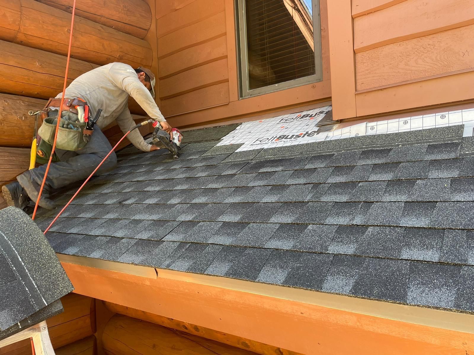 Roofer in tan shirt and pants, nailing shingles to a brown log cabin roof.