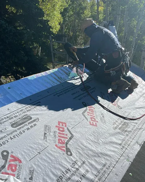 A construction worker kneels on a sloped roof covered in Epilay underlayment, using a nail gun to install roofing materials.