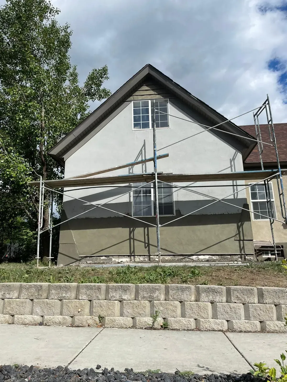 House under construction, scaffold, stucco exterior, retaining wall, cloudy sky.