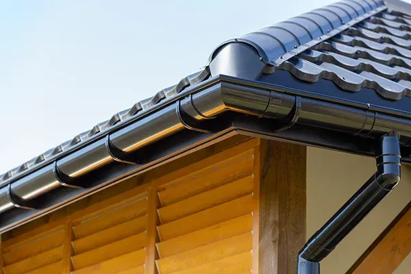 Black rain gutter on a roof's edge, visible wooden structure and blue sky.