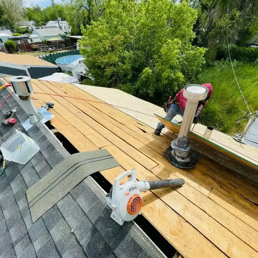 Roofer working on a roof, using a leaf blower. Partially shingled roof with wood planking visible.