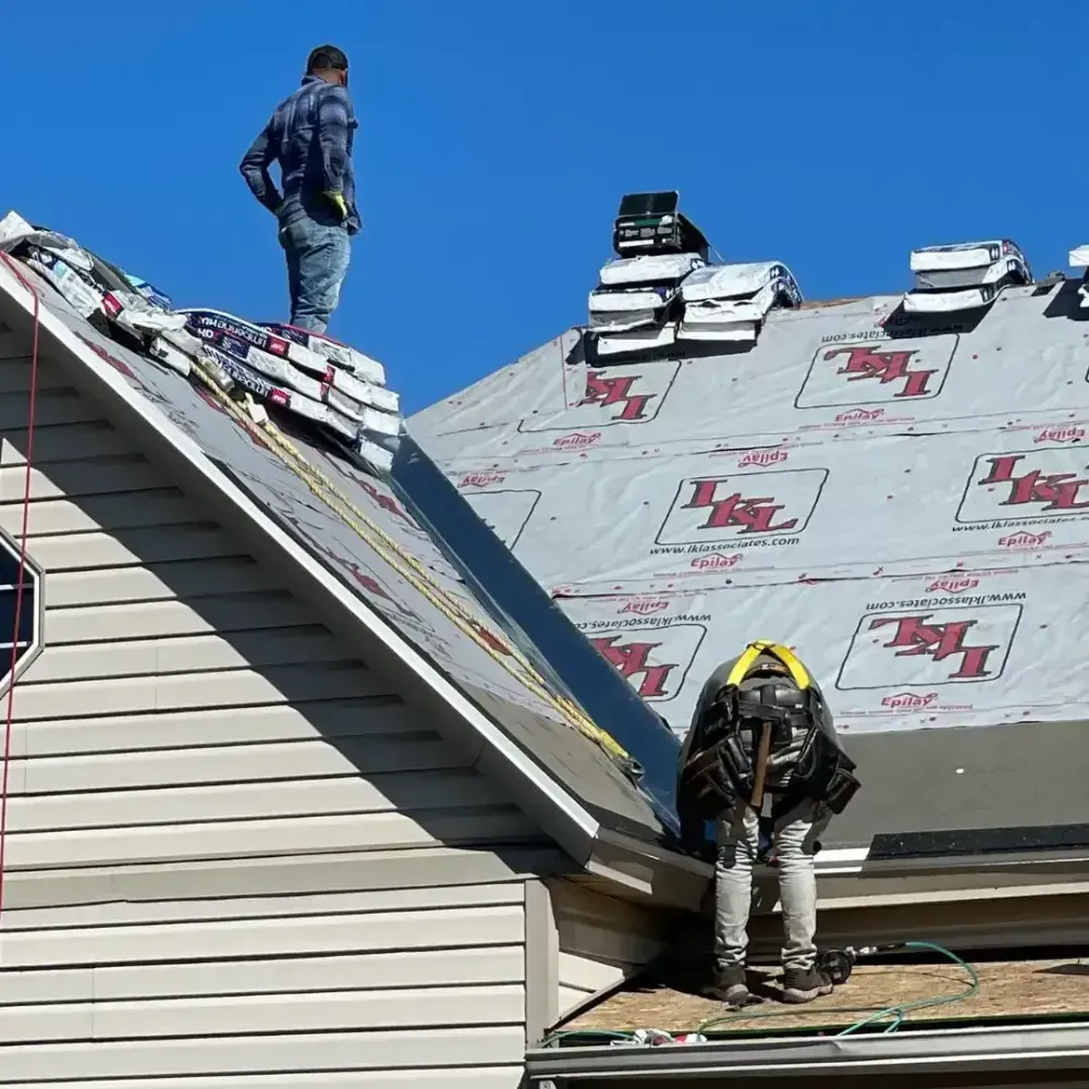 Two roofers working on a house roof. One stands, the other kneels, with safety harness. Blue sky.