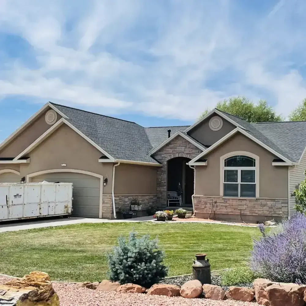 Tan stucco house with stone accents, gray roof, and green lawn. A dumpster is in the driveway.