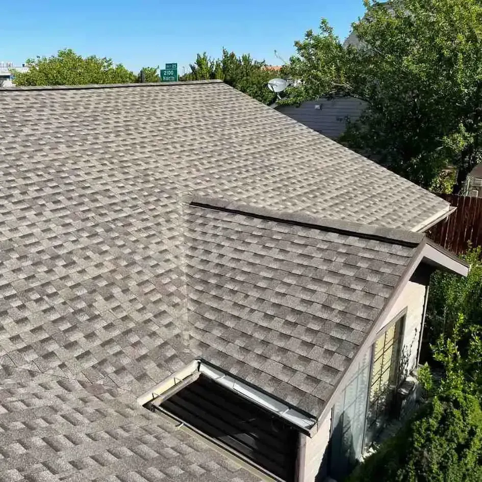 Gray asphalt shingle roof on a house, angled view. Sunny day, surrounded by trees.