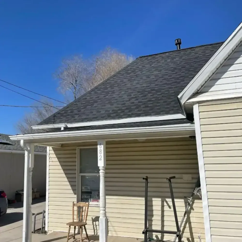 House with dark gray roof and tan siding, blue sky. Porch with chair.